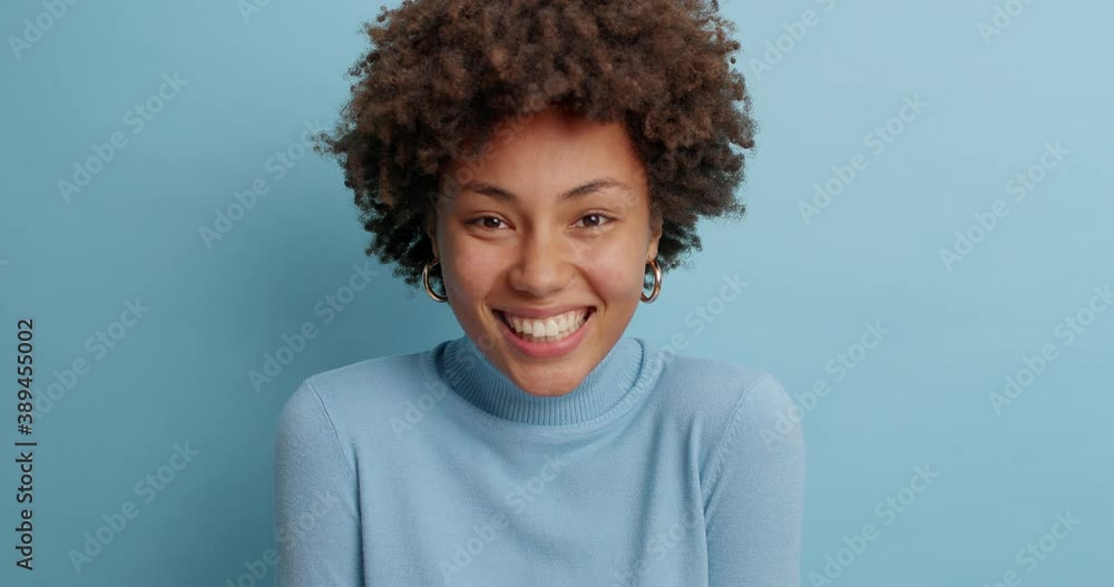 Cute positive dark skinned young woman with cheerful expression smiles gently expresses happiness enjoys pleasant talk dressed in casual poloneck isolated over blue background. Emotions concept Cute positive dark skinned young woman with cheerful expression smiles gently expresses happiness enjoys pleasant talk dressed in casual poloneck isolated over blue background. Emotions concept