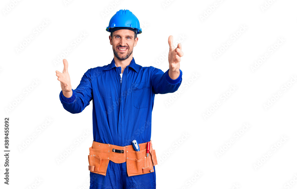 Young handsome man wearing worker uniform and hardhat looking at the camera smiling with open arms for hug. cheerful expression embracing happiness.