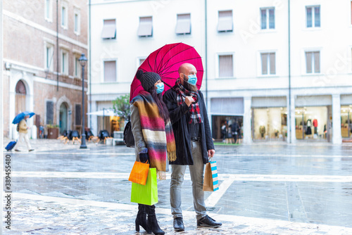 Beautiful couple with an umbrella and winter clothes is walking and smiling while doing shopping in city street on a storm rainy day. side view of a couple of a bearded balt man and brunette woman 