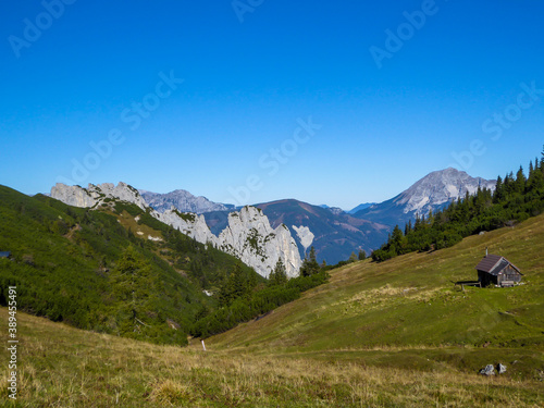 A panoramic view on the valley in Kaiserau Kreuzkogel region, Austrian Alps. There is a small cottage on the side of a meadow. Few trees on the slopes, endless mountain chains. Sunny and bright day.