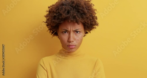 Beautiful dark skinned young woman looks angrily ar camera frowns face being irritated with something expresses negative emotions wears casual clothing isolated over yellow background. Annoyance