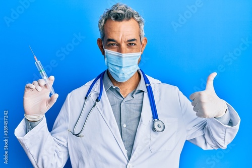 Middle age grey-haired man wearing doctor uniform and medical mask holding syringe smiling happy and positive, thumb up doing excellent and approval sign