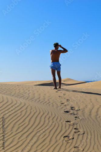Man in swimming trunks walking through the desert sand dunes.