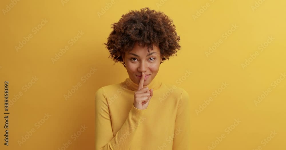 Happy mysterious woman with toothy smile puts index finger on lips has tricky gaze at camera tells secret only for you dressed in casual turtleneck isolated over yellow background spreads rumors Happy mysterious woman with toothy smile puts index finger on lips has tricky gaze at camera tells secret only for you dressed in casual turtleneck isolated over yellow background spreads rumors