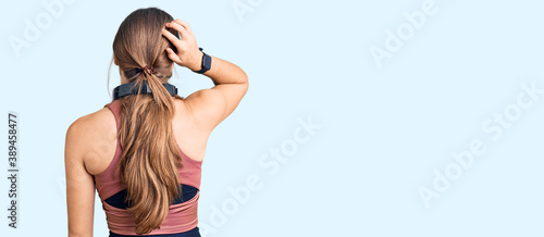 Beautiful caucasian young woman wearing gym clothes and using headphones backwards thinking about doubt with hand on head