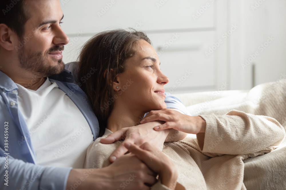 Warm embraces of love. Caring young spouses or couple in love spending free time at home together sitting on sofa embracing looking at distance enjoying each other company dreaming of happy future Warm embraces of love. Caring young spouses or couple in love spending free time at home together sitting on sofa embracing looking at distance enjoying each other company dreaming of happy future