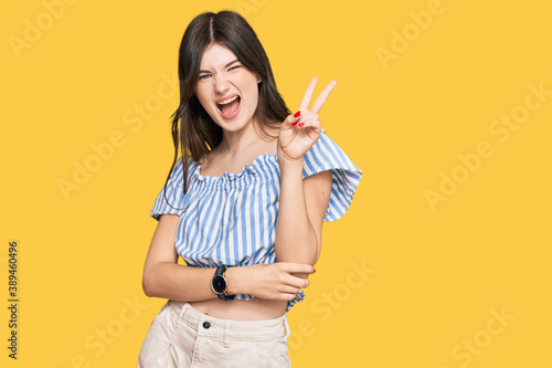 Young beautiful caucasian girl wearing casual clothes smiling with happy face winking at the camera doing victory sign with fingers. number two.