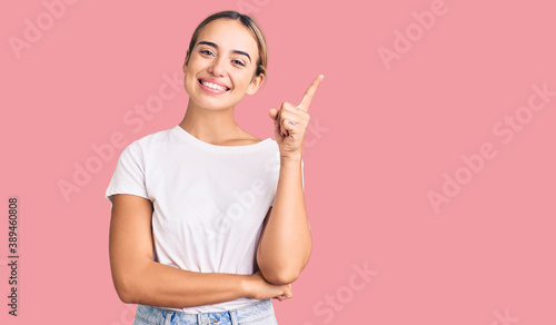 Young beautiful blonde woman wearing casual white tshirt with a big smile on face, pointing with hand and finger to the side looking at the camera. photo