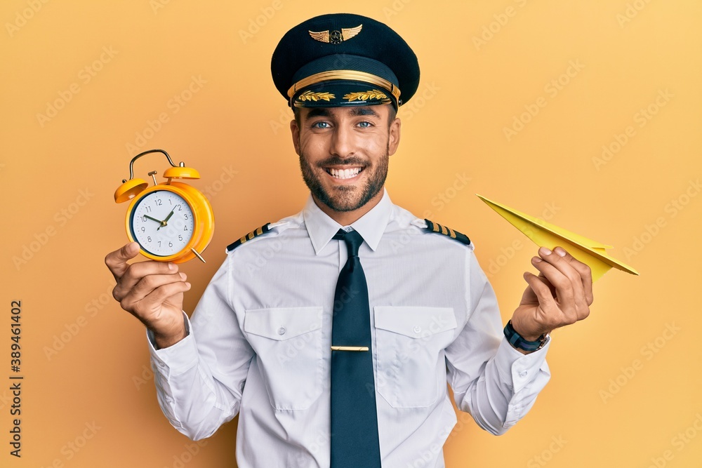 Handsome hispanic pilot man holding paper plane and alarm clock smiling