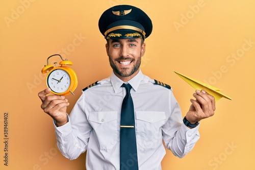 Valokuva Handsome hispanic pilot man holding paper plane and alarm clock smiling with a happy and cool smile on face