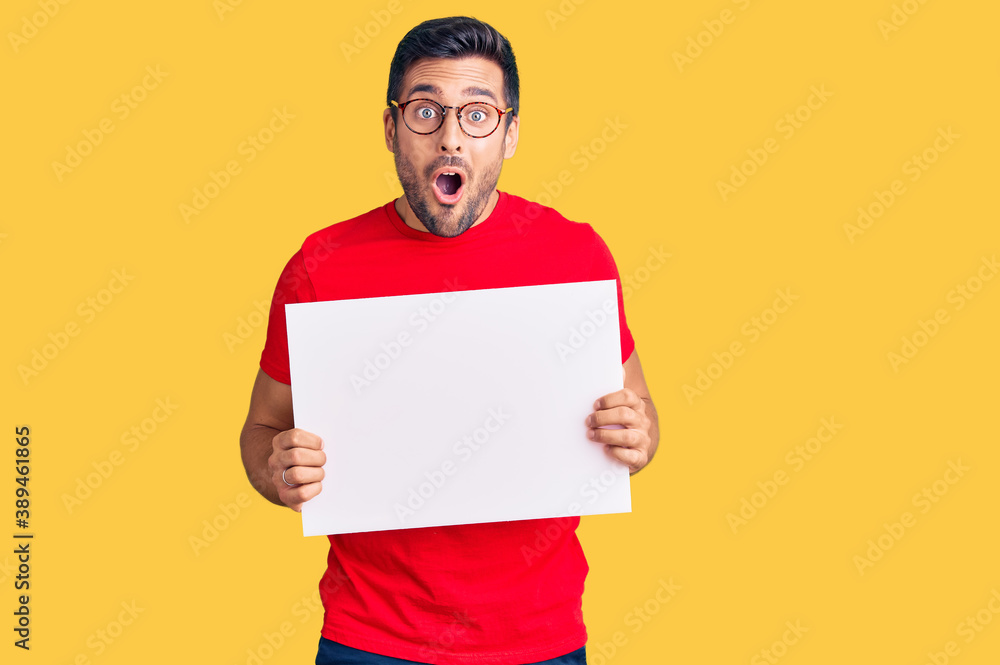 Young hispanic man holding blank empty banner scared and amazed with open mouth for surprise, disbelief face Young hispanic man holding blank empty banner scared and amazed with open mouth for surprise, disbelief face