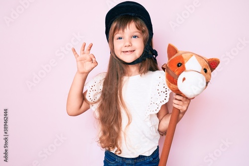 Little caucasian kid girl with long hair riding horse toy wearing vintage helmet doing ok sign with fingers, smiling friendly gesturing excellent symbol photo