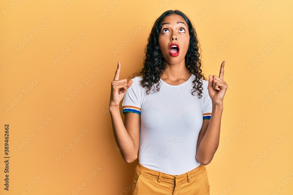 Young african american girl wearing casual clothes amazed and surprised looking up and pointing with fingers and raised arms. Young african american girl wearing casual clothes amazed and surprised looking up and pointing with fingers and raised arms.