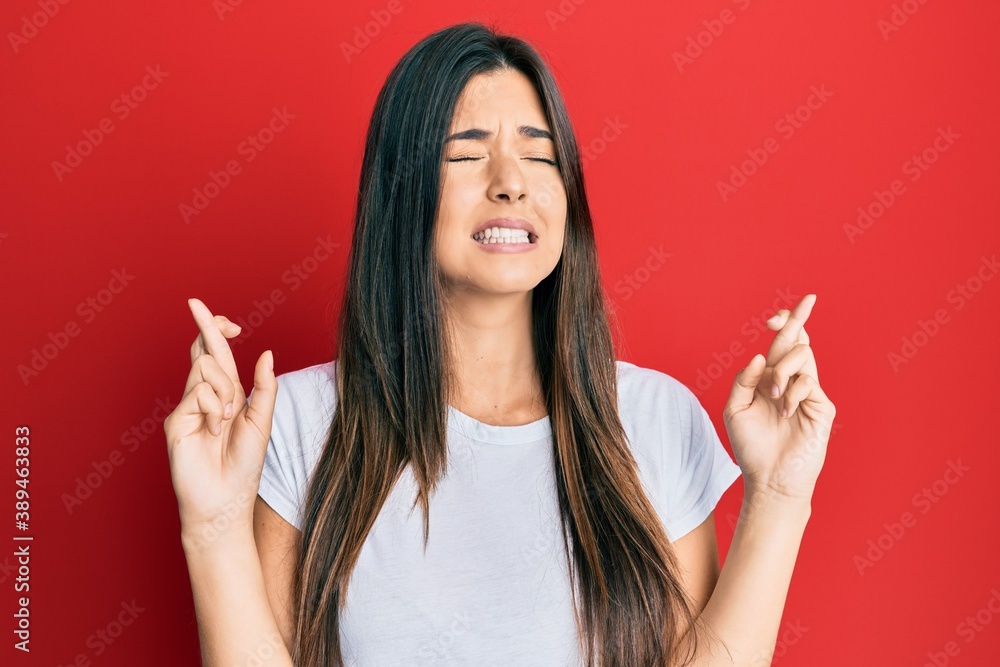 Young brunette woman wearing casual white tshirt over red background gesturing finger crossed smiling with hope and eyes closed. luck and superstitious concept. Young brunette woman wearing casual white tshirt over red background gesturing finger crossed smiling with hope and eyes closed. luck and superstitious concept.
