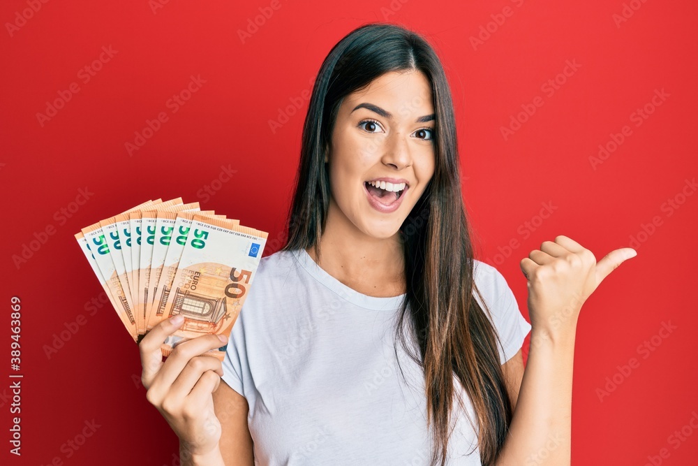 Young brunette woman holding 50 euro banknotes pointing thumb up to the side smiling happy with open mouth - obrazy, fototapety, plakaty 