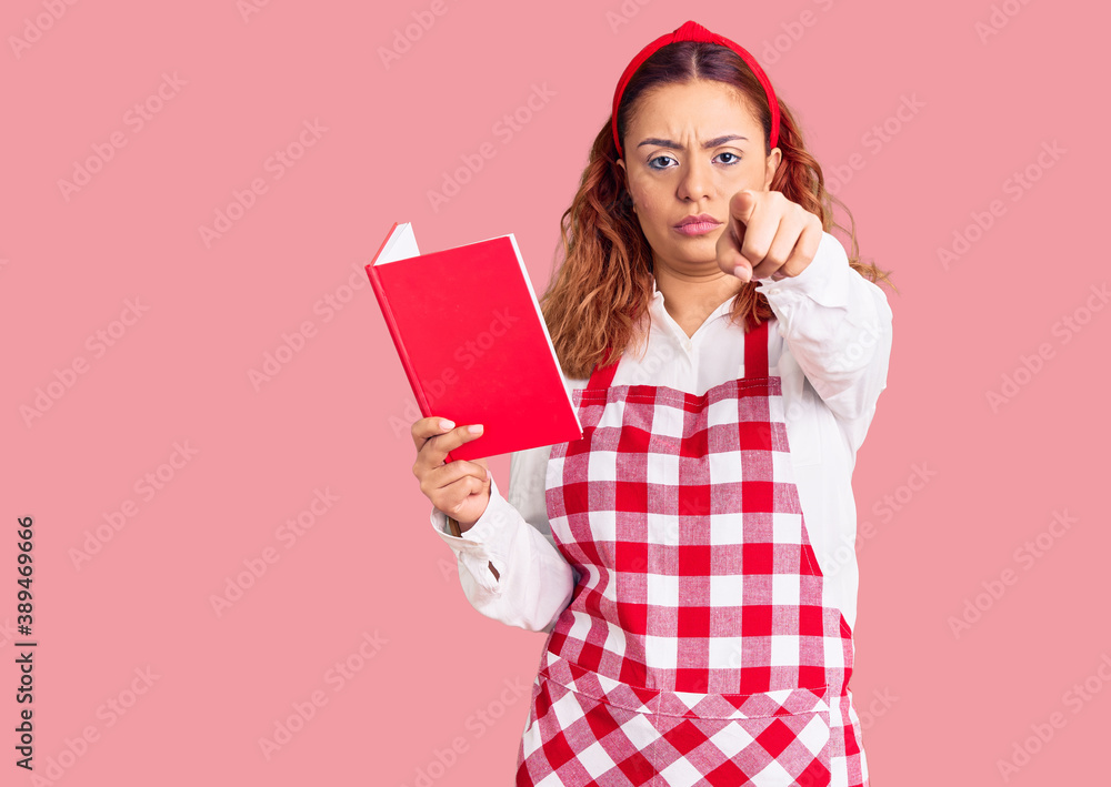 Young latin woman wearing apron holding recipe book pointing with finger to the camera and to you, confident gesture looking serious Young latin woman wearing apron holding recipe book pointing with finger to the camera and to you, confident gesture looking serious