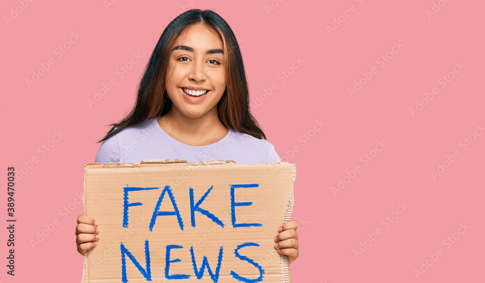 Young latin woman holding fake news banner looking positive and happy standing and smiling with a confident smile showing teeth Young latin woman holding fake news banner looking positive and happy standing and smiling with a confident smile showing teeth