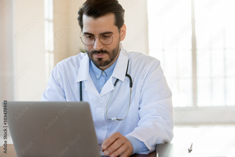 Thoughtful focused male physician working on laptop in hospital office counseling online, providing virtual appointment, studying new academic research by his speciality, thinking on electronic report
