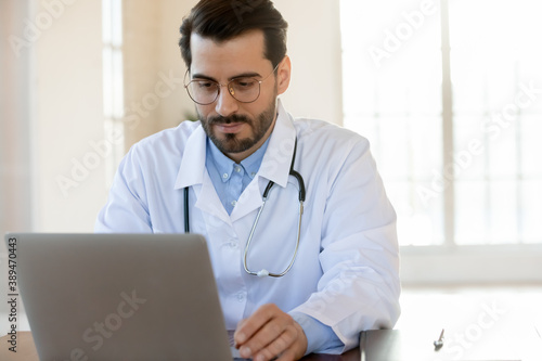 Thoughtful focused male physician working on laptop in hospital office counseling online  providing virtual appointment  studying new academic research by his speciality  thinking on electronic report