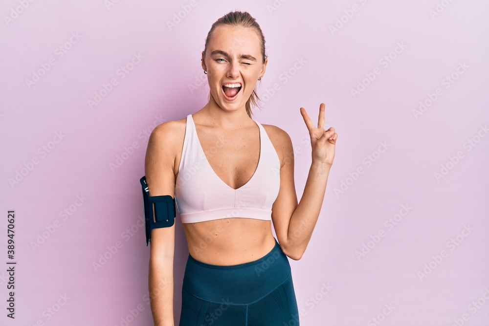 Beautiful blonde woman wearing sportswear and arm band smiling with happy face winking at the camera doing victory sign. number two.