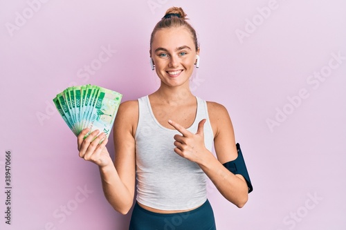 Beautiful blonde sport woman holding russian 100 ruble banknotes smiling happy pointing with hand and finger