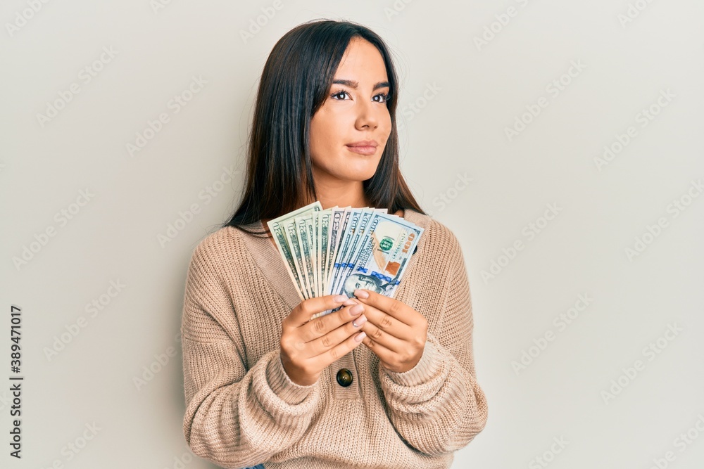 Young beautiful hispanic girl holding dollars smiling looking to the side and staring away thinking. Young beautiful hispanic girl holding dollars smiling looking to the side and staring away thinking.