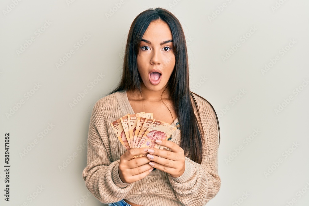 Young beautiful hispanic girl holding mexican pesos afraid and shocked with surprise and amazed expression, fear and excited face. Young beautiful hispanic girl holding mexican pesos afraid and shocked with surprise and amazed expression, fear and excited face.