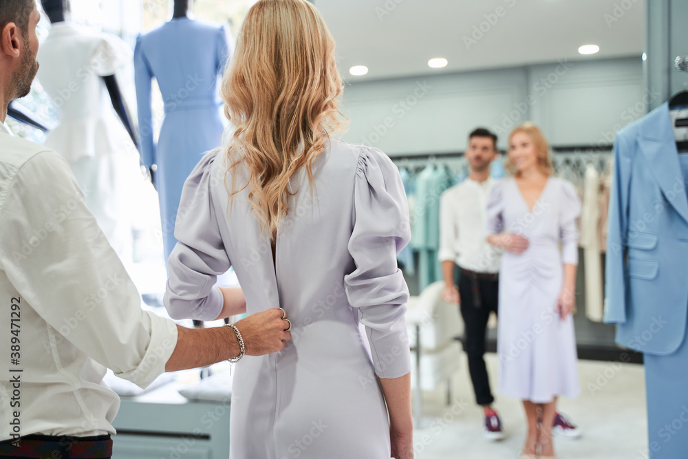 Woman looking at the mirror while stylist buttoning the dress Woman looking at the mirror while stylist buttoning the dress
