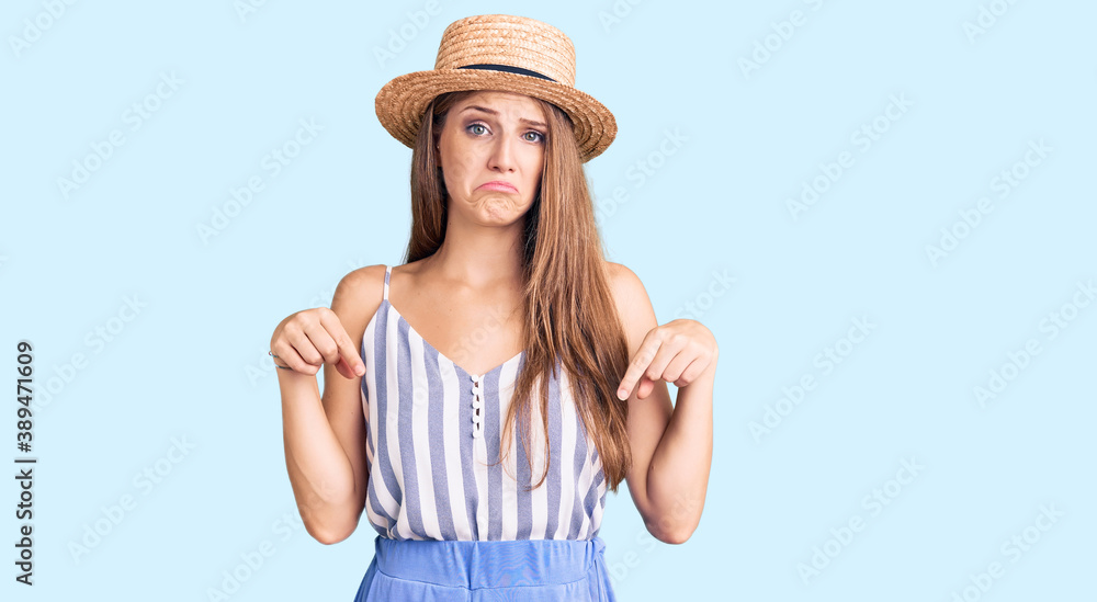 Young beautiful blonde woman wearing summer hat pointing down looking sad and upset, indicating direction with fingers, unhappy and depressed. Young beautiful blonde woman wearing summer hat pointing down looking sad and upset, indicating direction with fingers, unhappy and depressed.