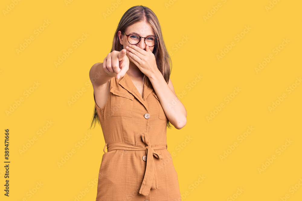Young beautiful blonde woman wearing casual clothes and glasses laughing at you, pointing finger to the camera with hand over mouth, shame expression Young beautiful blonde woman wearing casual clothes and glasses laughing at you, pointing finger to the camera with hand over mouth, shame expression