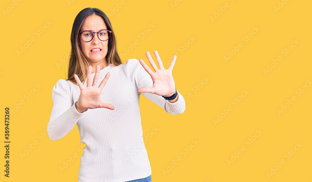 Beautiful brunette young woman wearing casual white sweater and glasses afraid and terrified with fear expression stop gesture with hands, shouting in shock. panic concept. Beautiful brunette young woman wearing casual white sweater and glasses afraid and terrified with fear expression stop gesture with hands, shouting in shock. panic concept.