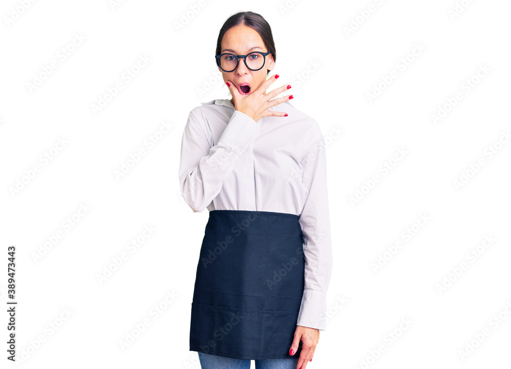 Beautiful brunette young woman wearing professional waitress apron looking fascinated with disbelief, surprise and amazed expression with hands on chin Beautiful brunette young woman wearing professional waitress apron looking fascinated with disbelief, surprise and amazed expression with hands on chin