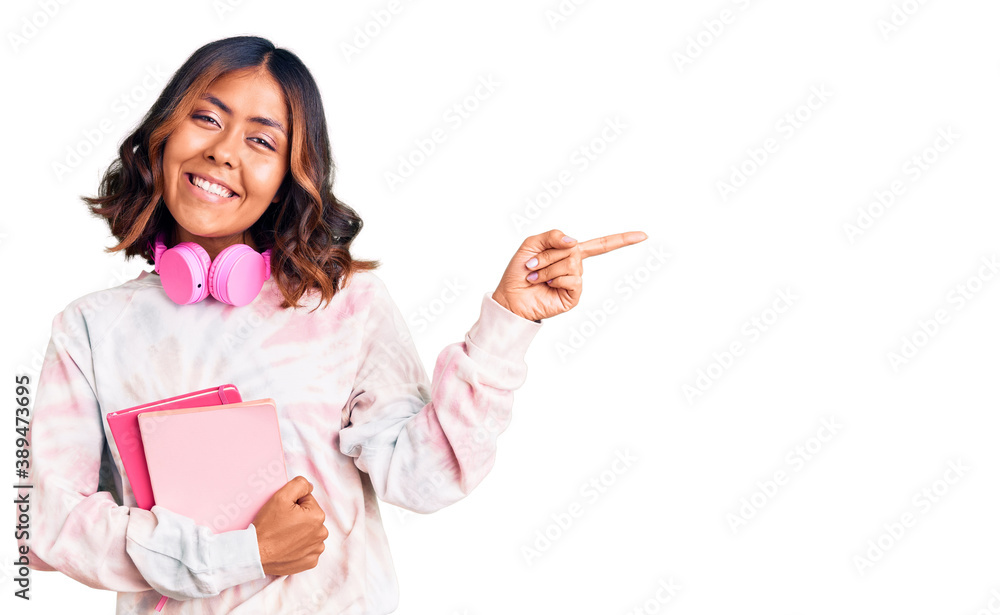 Young beautiful mixed race woman holding books wearing headphones smiling happy pointing with hand and finger to the side Young beautiful mixed race woman holding books wearing headphones smiling happy pointing with hand and finger to the side