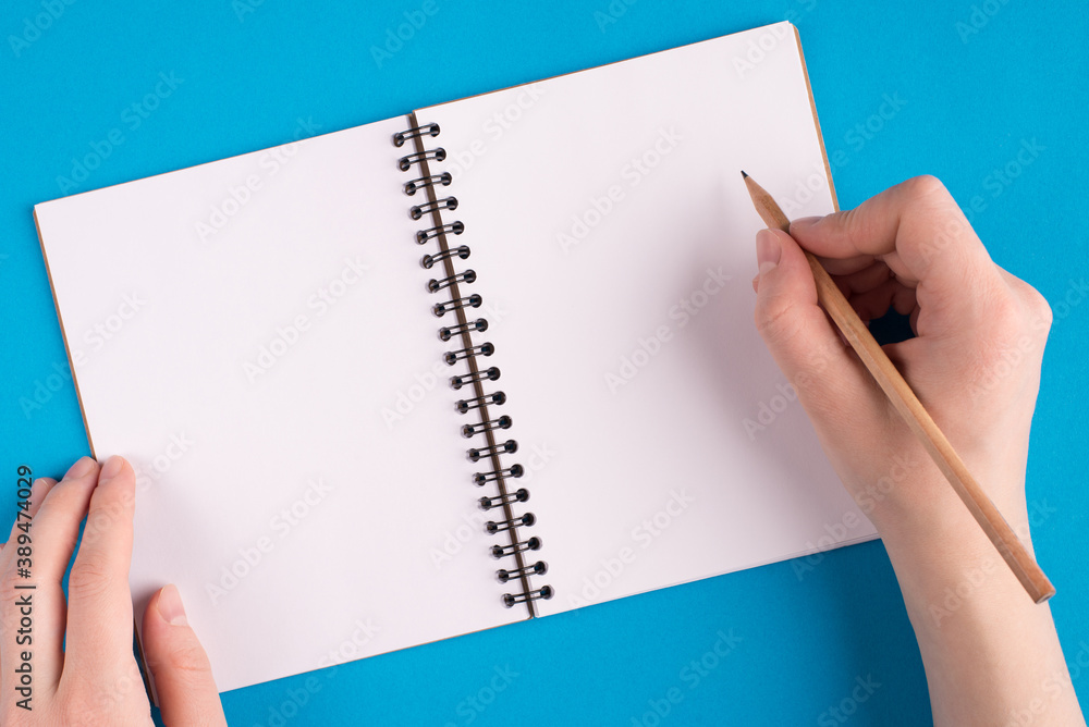 Start writing business start up concept. Top above overhead first person close up photo of female teen hipster beginning to write new ideas into notebook isolated bright blue background table desk Start writing business start up concept. Top above overhead first person close up photo of female teen hipster beginning to write new ideas into notebook isolated bright blue background table desk
