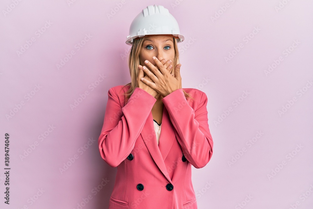 Beautiful middle age blonde woman wearing architect hardhat shocked covering mouth with hands for mistake. secret concept.