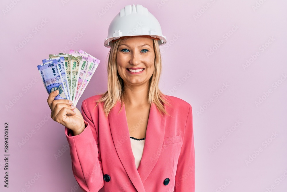 Beautiful middle age blonde woman wearing architect hardhat holding indian rupee looking positive and happy standing and smiling with a confident smile showing teeth Beautiful middle age blonde woman wearing architect hardhat holding indian rupee looking positive and happy standing and smiling with a confident smile showing teeth