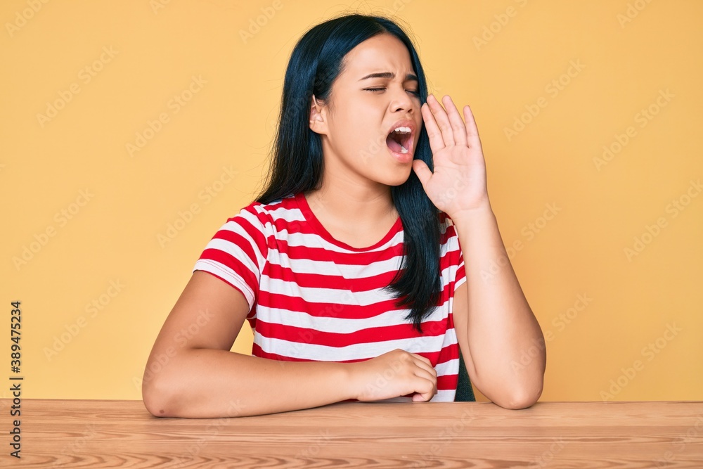 Young beautiful asian girl wearing casual clothes sitting on the table shouting and screaming loud to side with hand on mouth. communication concept. Young beautiful asian girl wearing casual clothes sitting on the table shouting and screaming loud to side with hand on mouth. communication concept.