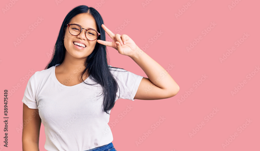 Young beautiful asian girl wearing casual clothes and glasses doing peace symbol with fingers over face, smiling cheerful showing victory Young beautiful asian girl wearing casual clothes and glasses doing peace symbol with fingers over face, smiling cheerful showing victory