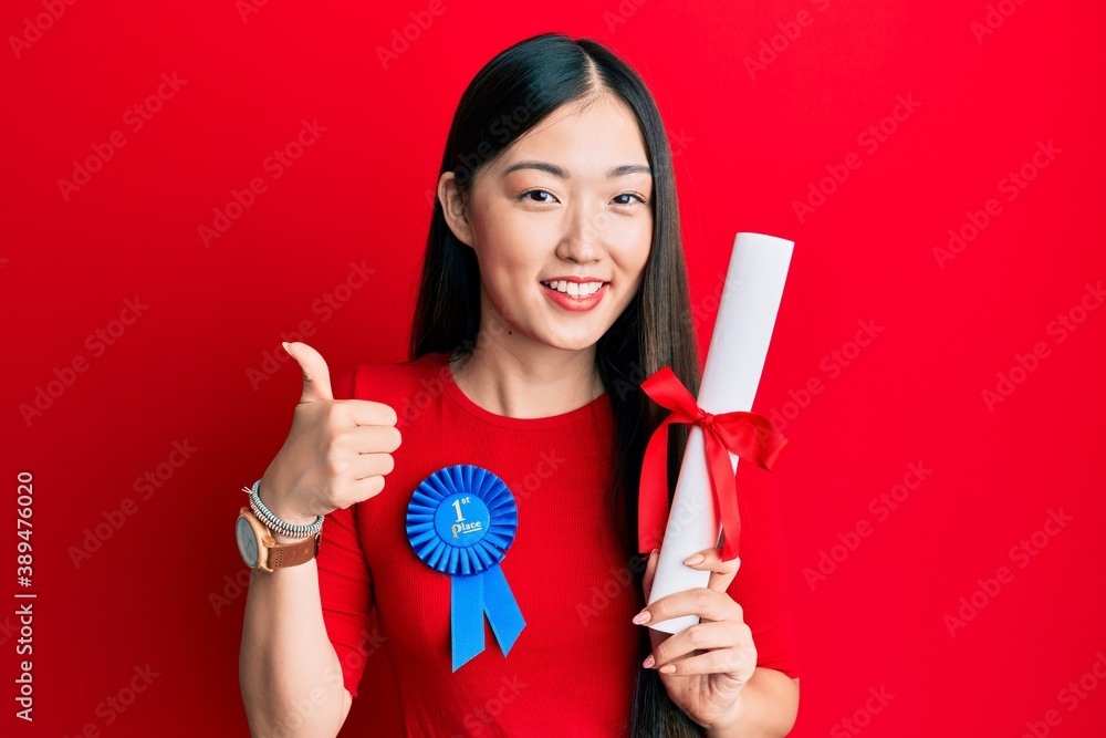 Young chinese woman wearing first place badge holding diploma smiling happy and positive, thumb up doing excellent and approval sign Young chinese woman wearing first place badge holding diploma smiling happy and positive, thumb up doing excellent and approval sign