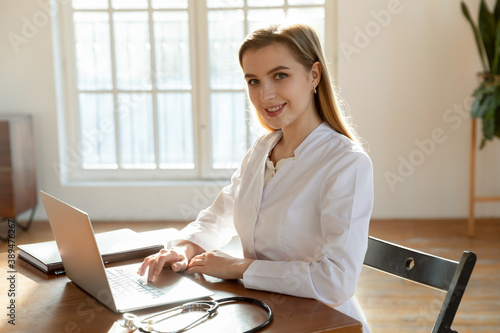 Welcome to our clinic. Portrait of pleasant confident young lady doctor, nurse or medical registrar sitting at workplace in office at modern hospital using laptop looking at camera with friendly smile