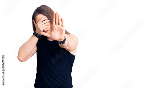 Young adult man with long hair wearing goth style with black clothes covering eyes with hands and doing stop gesture with sad and fear expression. embarrassed and negative concept.