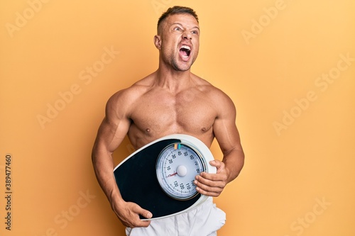 Handsome muscle man holding weight machine to balance weight loss angry and mad screaming frustrated and furious  shouting with anger looking up.
