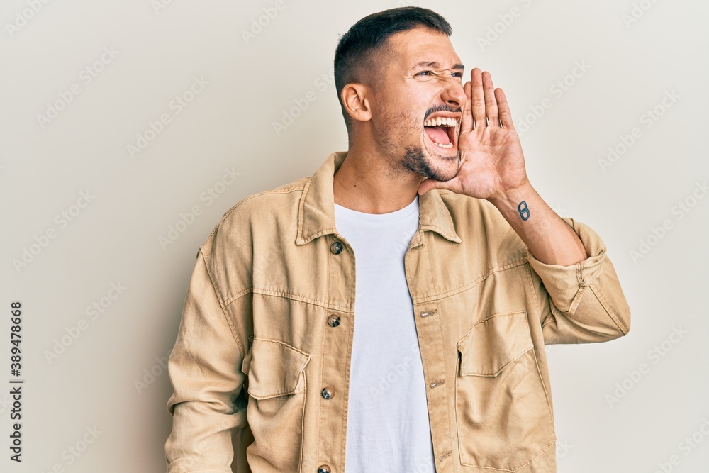 Handsome man with tattoos wearing casual brown denim jacket shouting and screaming loud to side with hand on mouth. communication concept. Handsome man with tattoos wearing casual brown denim jacket shouting and screaming loud to side with hand on mouth. communication concept.