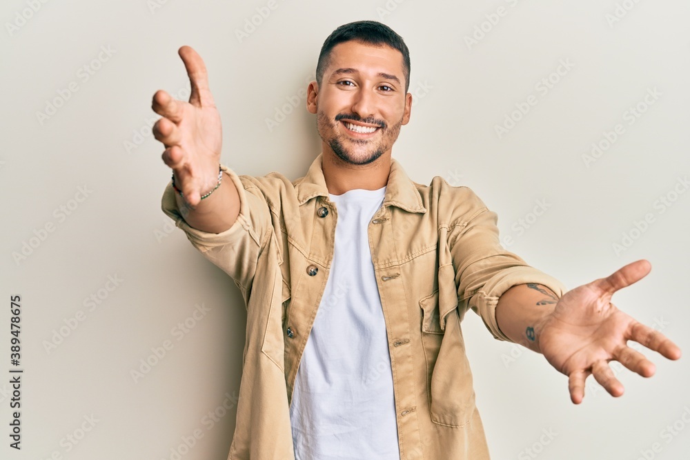 Handsome man with tattoos wearing casual brown denim jacket looking at the camera smiling with open arms for hug. cheerful expression embracing happiness. Handsome man with tattoos wearing casual brown denim jacket looking at the camera smiling with open arms for hug. cheerful expression embracing happiness.