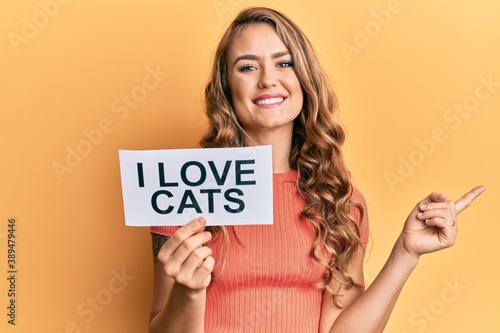 Young blonde girl holding paper with i love cats phrase smiling happy pointing with hand and finger to the side