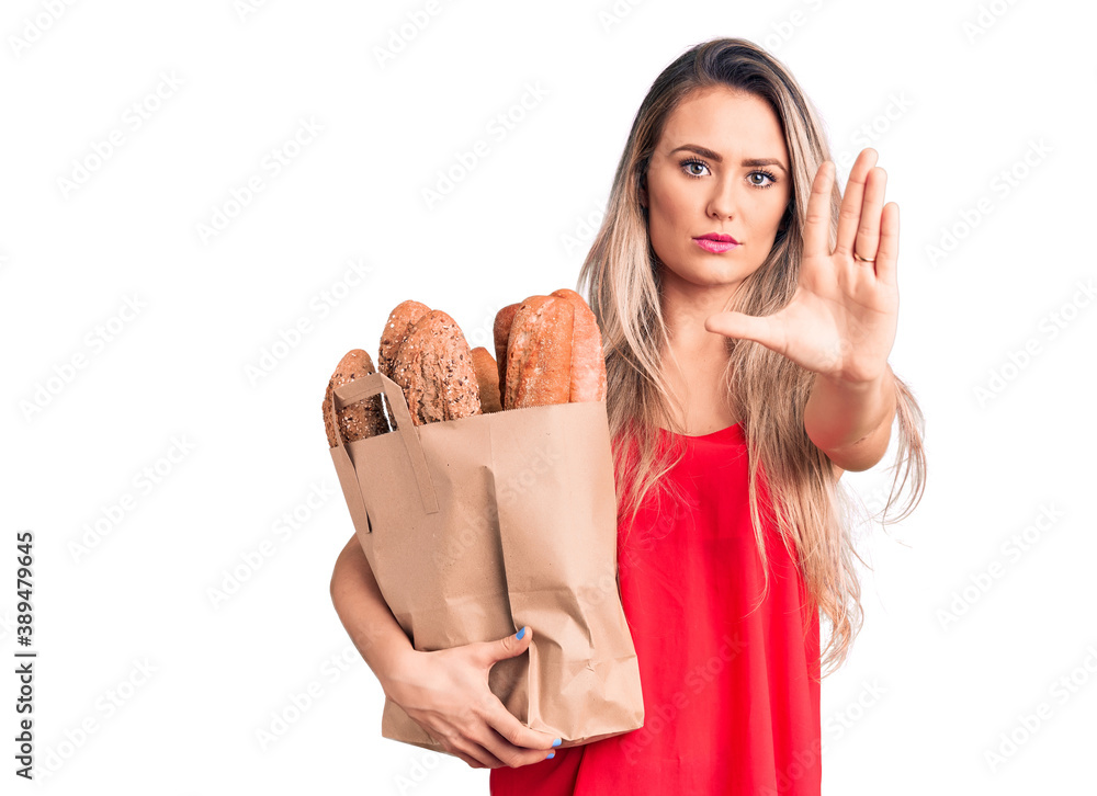 Young beautiful blonde woman holding paper bag with bread with open hand doing stop sign with serious and confident expression, defense gesture Young beautiful blonde woman holding paper bag with bread with open hand doing stop sign with serious and confident expression, defense gesture
