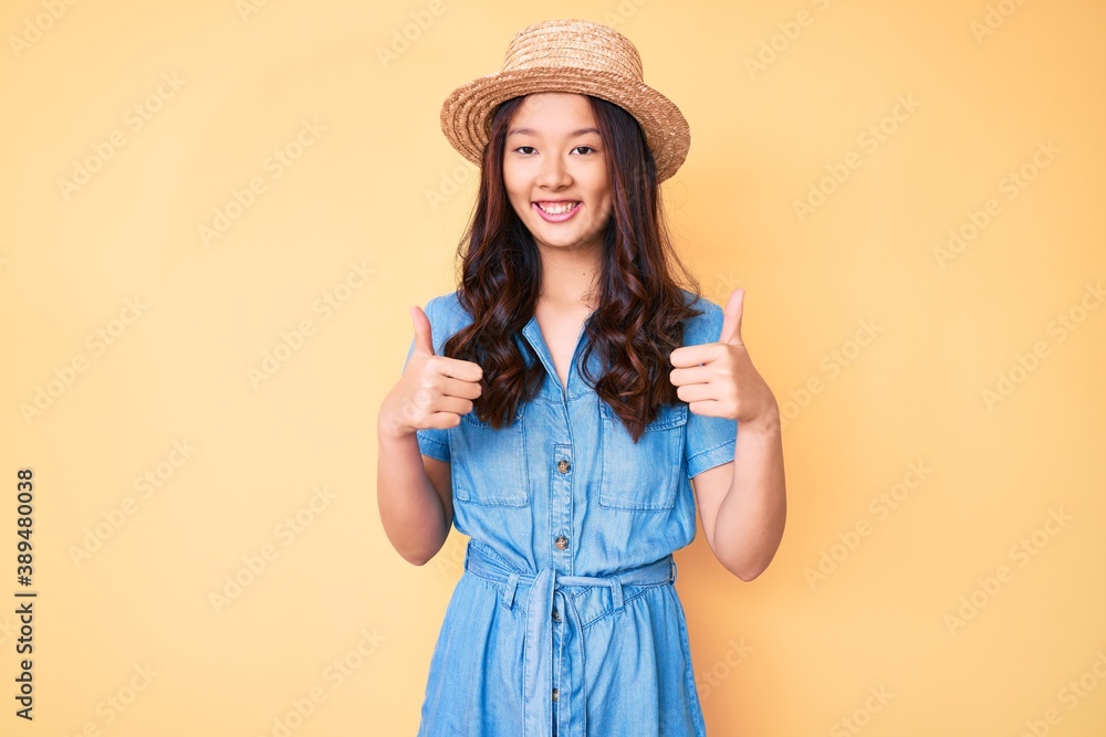 Young beautiful chinese girl wearing summer hat success sign doing positive gesture with hand, thumbs up smiling and happy. cheerful expression and winner gesture. Young beautiful chinese girl wearing summer hat success sign doing positive gesture with hand, thumbs up smiling and happy. cheerful expression and winner gesture.