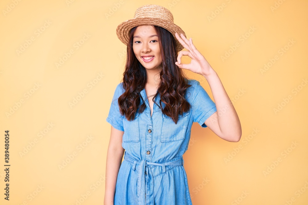 Young beautiful chinese girl wearing summer hat smiling positive doing ok sign with hand and fingers. successful expression. Young beautiful chinese girl wearing summer hat smiling positive doing ok sign with hand and fingers. successful expression.