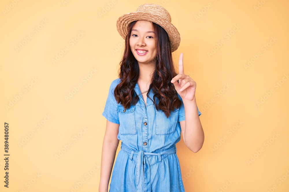 Young beautiful chinese girl wearing summer hat showing and pointing up with finger number one while smiling confident and happy. Young beautiful chinese girl wearing summer hat showing and pointing up with finger number one while smiling confident and happy.