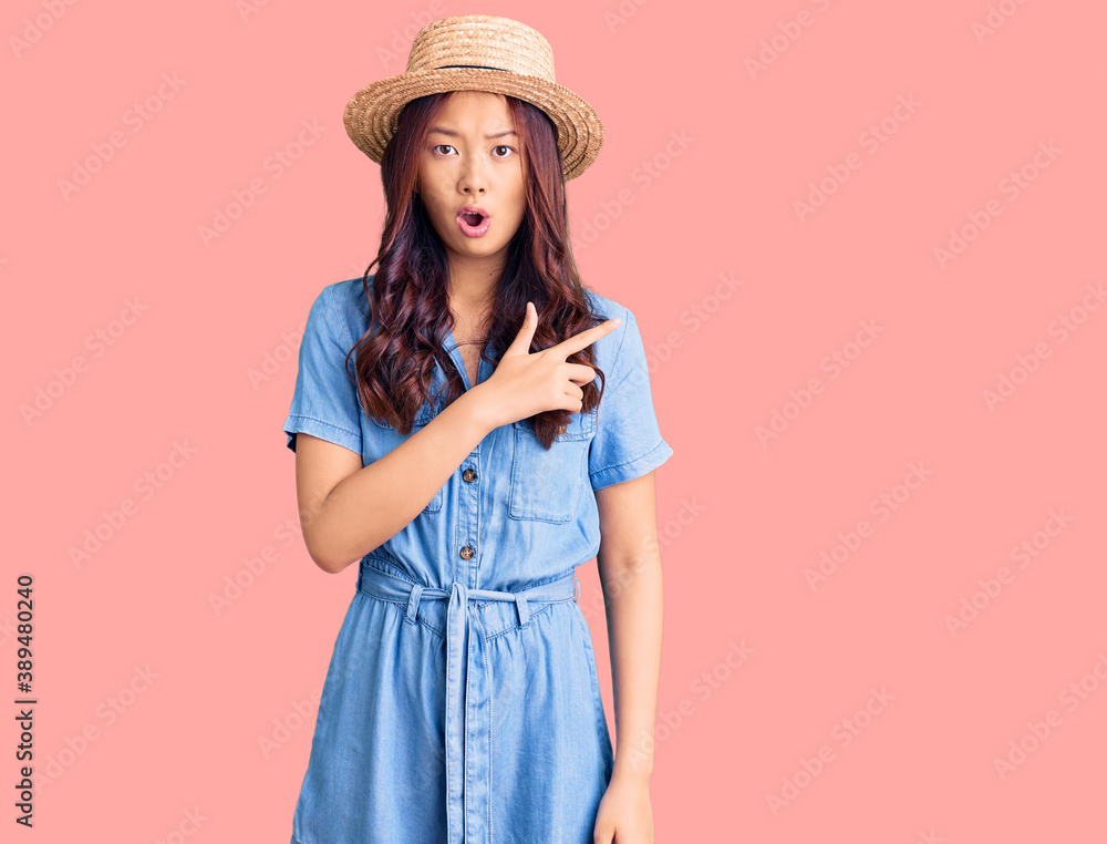 Young beautiful chinese girl wearing summer hat surprised pointing with finger to the side, open mouth amazed expression. Young beautiful chinese girl wearing summer hat surprised pointing with finger to the side, open mouth amazed expression.